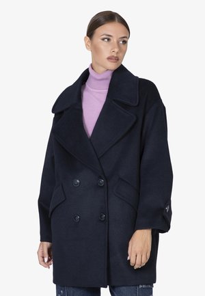 Donkerblauw dubbelbreasted jas van een wolmix. Kenmerken zijn grote revers, twee voorkledingzakken en zwarte knopen. Het model draagt een lichtpaarse coltrui.