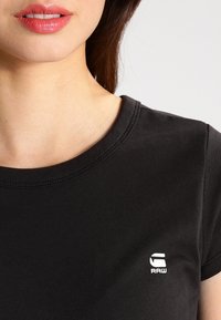 Svart bomullströja med rund halsringning, med en liten vit "RAW"-logotyp på vänster sida. Slät textur och figurnära design.