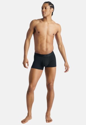 Atletische man met gevlochten haar staat blootsvoets in zwarte boxershorts en kijkt naar de zijkant tegen een eenvoudige lichte achtergrond.