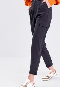 Person iført sorte cargo bukser med sidelommer og hvide sneakers med pink og brune accenter, stående på en ensfarvet hvid baggrund.