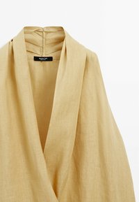 Haut enveloppant en lin beige avec un décolleté en V et un drapé doux. Détails plissés à l'arrière et étiquette de marque à l'intérieur du décolleté.