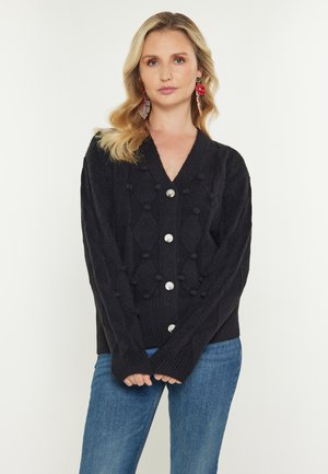 Frau mit blondem Haar, die eine schwarze, strukturierte Strickjacke mit Knöpfen und blaue Jeans trägt, vor einem schlichten hellen Hintergrund.