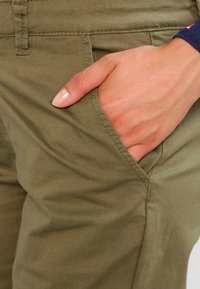 Lekkie spodnie z tkaniny w kolorze oliwkowym, z bocznymi kieszeniami, gładką strukturą i widocznymi szwami wzdłuż krawędzi.