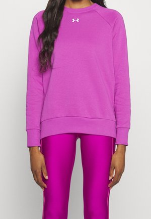 Kobieta w bluzie Under Armour z długim rękawem w kolorze magenta i błyszczących magentowych legginsach, stojąca na tle jasnego, jednolitego tła.