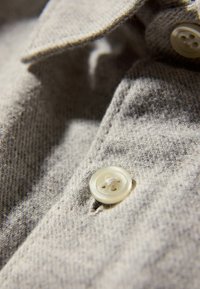 Lichtgrijze stof met een fijne textuur, met een close-up van een knoop met vier gaten, genaaid naast een kraag.