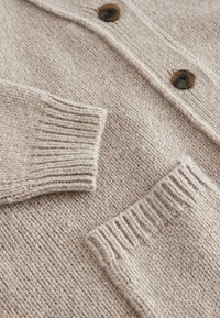 Tissu de cardigan en maille beige avec poignets côtelés, poche plaquée à l'avant et boutons marron marbrés le long de la patte de boutonnage.