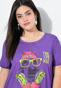 T-shirt graphique violet avec un dessin animé de chien portant des écouteurs, des lunettes de soleil vert fluo et un chapeau rose avec le texte "FAIS DU BRUIT."