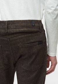 Άτομο που φορά σκούρο καφέ τζιν denim και λευκό πουκάμισο με μακριά μανίκια, δείχνοντας τις πίσω τσέπες και μια μικρή ετικέτα με το λογότυπο στη μέση.