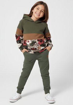 Uśmiechnięte dziecko stoi z rękami w kieszeniach, ubrane w zielone spodnie, białe trampki i zielono-brązową bluzę z kwiatowym wzorem.