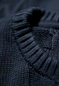 Marinefarbener Strickstoff mit geripptem Textur am Halsauschnitt; Nahaufnahme zeigt kunstvolle Webmuster und verschiedene Sticharten.