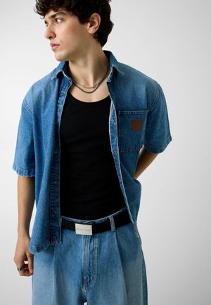Jonge man in gelaagde denim outfit met een shirt met korte mouwen, een zwart hemdje, zilveren kettingen en losse jeans, kijkend naar links tegen een effen achtergrond.