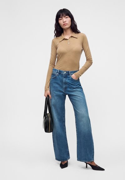 Femme portant un polo beige à manches longues, jean bleu à jambes larges, talons pointus noirs, tenant un sac à main noir sur un fond uni.