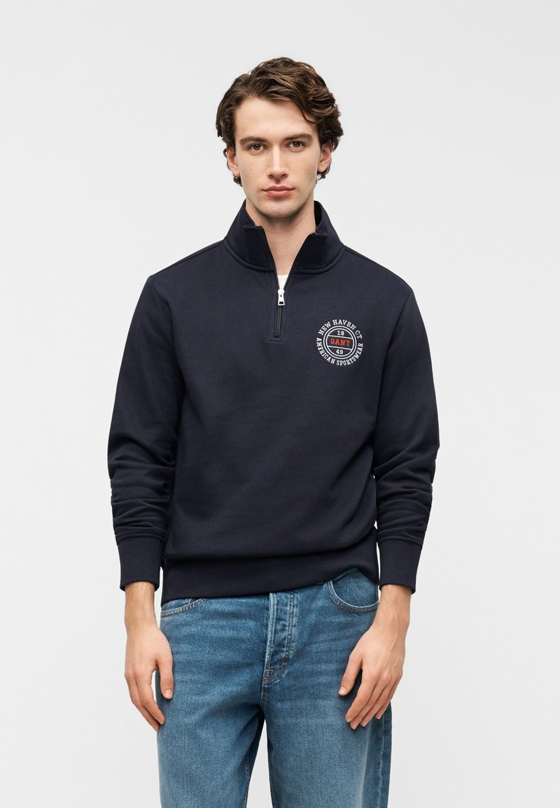 Felpa blu navy con mezza zip e collo alto, con un logo circolare stampato sul petto. Indossata con jeans azzurri.