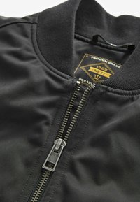 Veste bomber noire dotée d'une fermeture éclair à l'avant, d'un col côtelé et d'une texture lisse. L'étiquette intérieure indique la marque et des informations sur la durabilité.