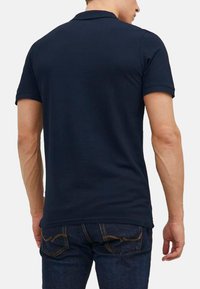 Marineblaues Polo-Shirt mit kurzen Ärmeln, einem Kragen und seitlichen Schlitzen. Getragen mit dunklen Jeans, die ein enges Design und eine glatte Textur zeigen.