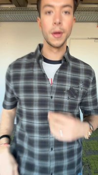 Νέος άνδρας που φοράει μαύρο και λευκό καρό πουκάμισο με κεντημένα αστέρια, στέκεται σε εσωτερικό χώρο μπροστά σε λευκό τοίχο και πόρτα.