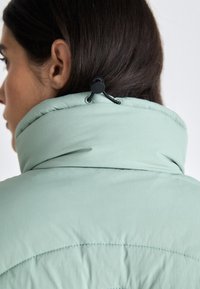 Collo del giubbotto imbottito verde menta con una texture morbida e un toggle nero regolabile per la vestibilità. Le cuciture visibili aggiungono dettaglio.