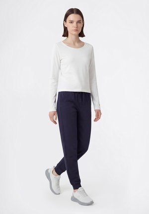 Donna con capelli castani lisci che indossa una maglia bianca a maniche lunghe, pantaloni jogger blu navy e scarpe da ginnastica bianche, in piedi davanti a uno sfondo bianco.