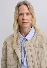 Chaqueta acolchada beige con un patrón curvado y cremallera plateada, usada sobre una camisa de rayas azul claro con cuello de botón.