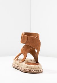 Bruna mockaskinnsandaler med ankelrem och vävd espadrille-yttersula samt öppen tå, uppställda mot en enkel vit bakgrund.