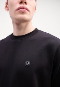 Sort sweatshirt lavet af glat stof, med rund hals og et lille cirkulært logo på brystet. Simpelt design uden mønstre.