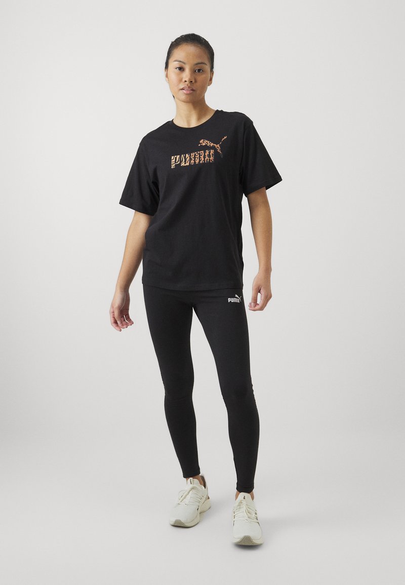 Camiseta de algodón negra con un logo de estampado de leopardo, combinada con leggings negros y zapatillas blancas. El conjunto presenta un corte holgado y un diseño sencillo.