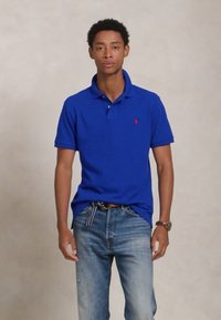 Mlad moški stoji, oblečen v modro polo majico in bledo modre kavbojke s črtastim pasom, ob bež ozadju.