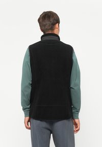 Zwarte fleece vest met hoge kraag, textuur aan de buitenkant en geen zichtbare sluitingen. Draag over een groen overhemd met lange mouwen, achteraanzicht.