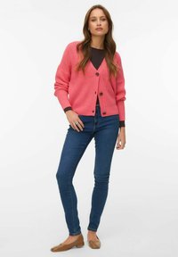 Mlada žena stoji, nosi ružičasti kardigan na kopčanje preko tamne majice, plave uske traperice i smeđe ravne cipele, na bijeloj pozadini.