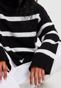 Gestreifter Pullover in Schwarz und Weiß mit einem hohen Kragen und breiten gerippten Bündchen. Die Textur wirkt gestrickt und weich. Trägt man mit weißen Hosen.