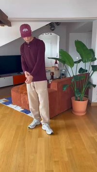 Camisa de manga larga burdeos, pantalones de chándal beige y zapatillas grises. Ambiente interior con suelo de madera, plantas y un sofá al fondo.