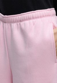 Gros plan sur un pantalon de survêtement à taille élastique rose clair porté avec un haut noir, montrant la poche latérale et une partie de la main dans la poche.