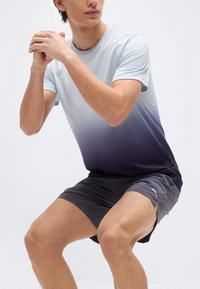 Άνδρας με μπλουζάκι σε αποχρώσεις μπλε-μοβ και γκρι σορτς Puma κάνει καθίσματα με τα χέρια ενωμένα μπροστά, πάνω σε λευκό φόντο.