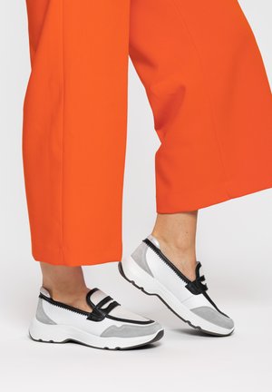 Persoon die feloranje wijde broek draagt en witte, grijze en zwarte instapsneakers met dikke zolen, staand met één voet omhoog.