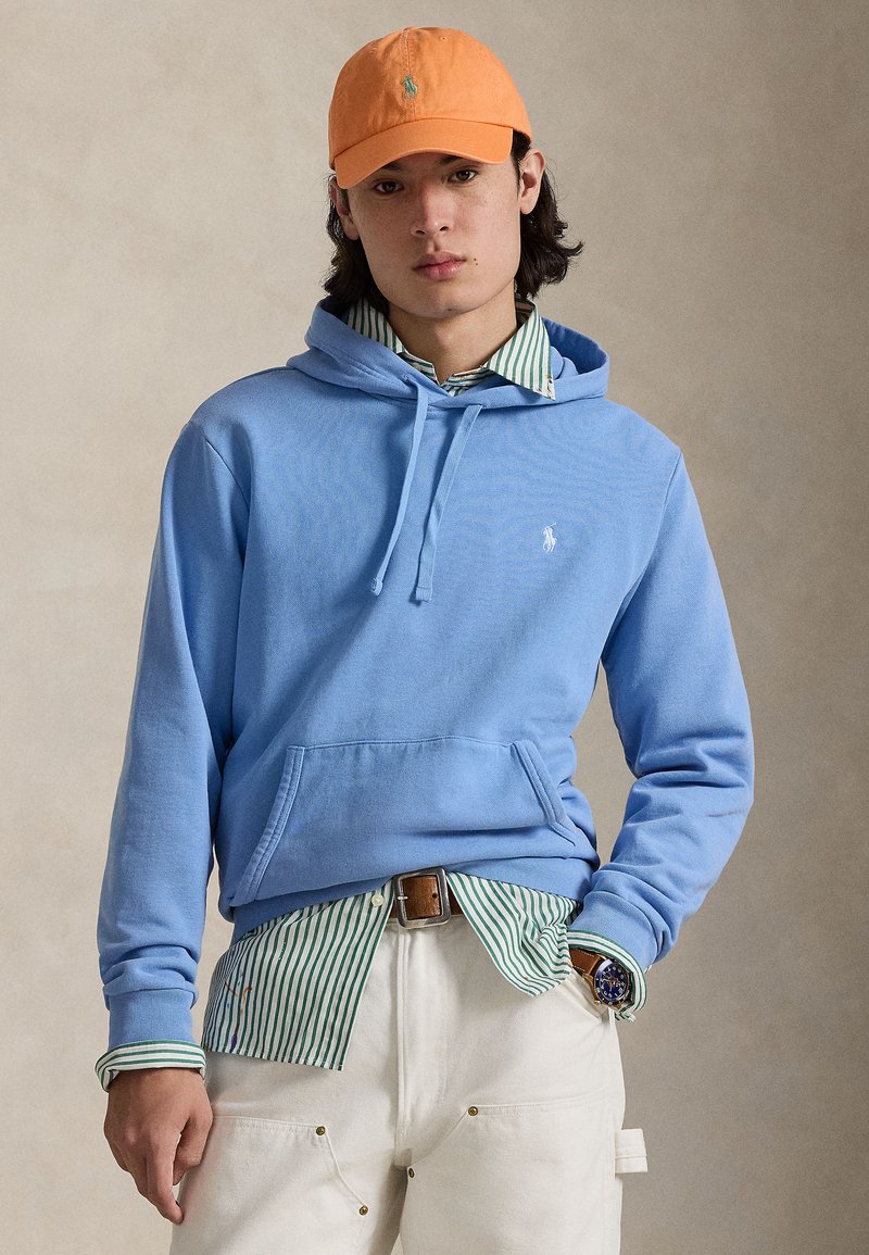 Φωτεινό γαλάζιο βαμβακερό φούτερ με κουκούλα, με τσέπη μπροστά και κορδόνι. Φορεμένο πάνω από πράσινο και λευκό ριγέ πουκάμισο με κουμπιά και λευκό παντελόνι. Πορτοκαλί καπέλο.