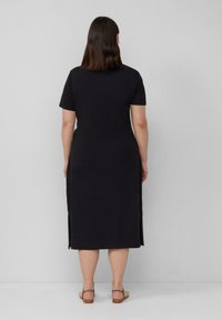 Schwarzes T-Shirt-Kleid mit kurzen Ärmeln und seitlichen Schlitzen. Glatte Textur und knielange Länge. Getragen mit hellbraunen Sandalen.