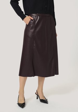 Kvinde iført mørkebrun læder midi-nederdel med sidelommer, sort cardigan og sorte slingback hæle står foran en lys baggrund.