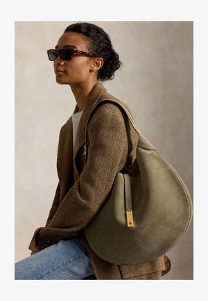 Femme portant des lunettes de soleil écaille et un blazer marron à chevrons, portant un grand sac à bandoulière en daim vert olive avec des ferrures dorées.