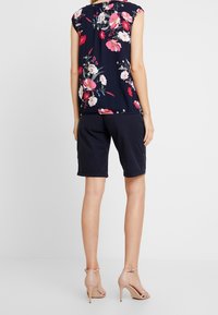 Marinblå blus med blommönster och korta ärmar, rosa och vita blommor, kombinerad med mörka shorts och beige högklackade sandaler.