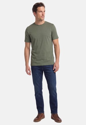 Mand stående iført en olivengrøn kortærmet t-shirt, mørkeblå jeans og brune snørestøvler, kigger mod venstre.