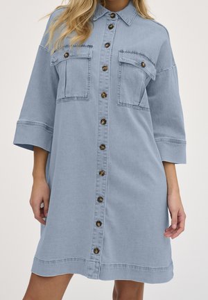 Lyseblå denimkjole med knapper, to brystlommer og trekvartlange ærmer, båret af en person med blondt hår.