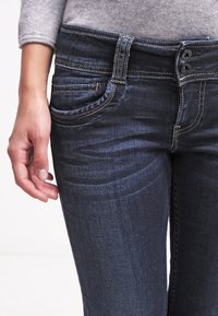 Pantalones vaqueros de mezclilla azul oscuro con cintura ajustada, cierre de dos botones y bolsillos traseros. Presentan un ligero desgastado y detalles de costura en contraste.