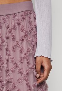 Lila blommig kjol med texturerade, broderade rosettvappliker. Mjukt, genomskinligt material med en bred midjeband. Lätt samlad design.