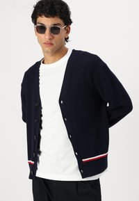 Mørkeblå strikket cardigan med v-hals, seks sorte knapper og en rød og hvit stripe nederst. Brukt over en hvit t-skjorte.