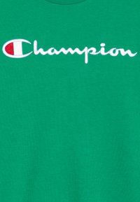 Grønn bomull t-skjorte med et fremtredende hvitt brodert "Champion"-logo og et lite rødt og hvitt emblem til venstre.