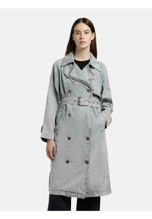 Donna che indossa un lungo trench grigio doppiopetto con cintura, in piedi con una mano in tasca davanti a uno sfondo semplice.