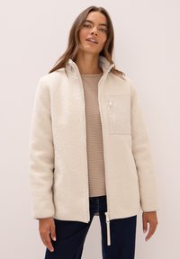 Veste en polaire couleur crème avec fermeture éclair sur le devant, col haut et poche en tissu contrastant. Portée par-dessus un haut à rayures à manches longues.