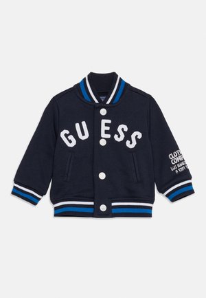 Veste universitaire pour enfants bleu marine avec boutons blancs, inscription "GUESS" sur le devant, et poignets et col rayés bleu et blanc.