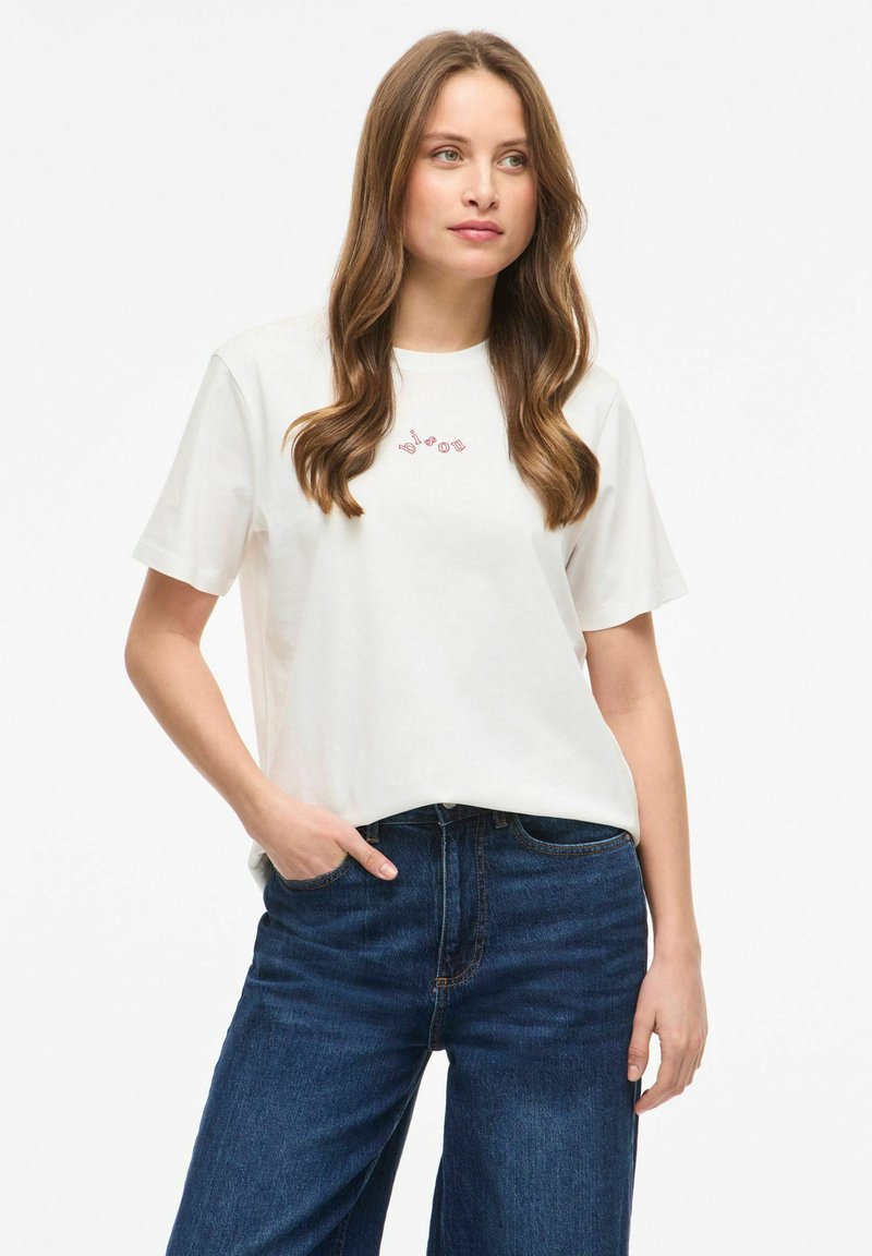 Jeune femme aux longs cheveux bruns portant un t-shirt blanc à manches courtes et un jean bleu foncé, debout avec une main dans la poche.