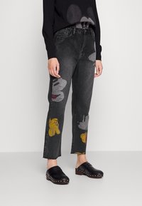 Jean en denim noir avec des motifs de mains de dessin animé colorées, jambes droites, ourlets effilochés et taille mi-haute. Associé à des sabots noirs.